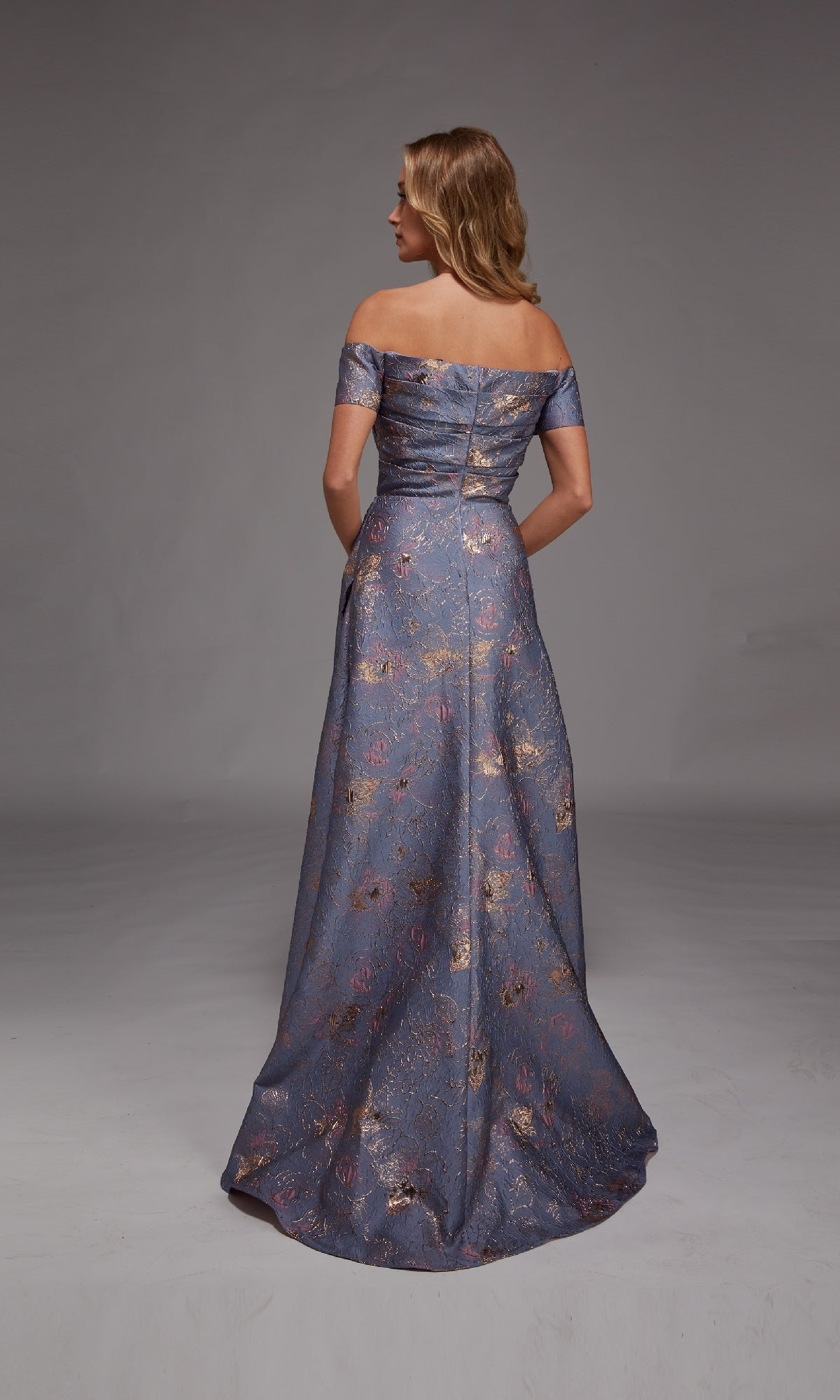 A woman stands with her back to the camera against a plain gray background, wearing the Alyce 27828 long formal dress—an off-the-shoulder, floor-length blue jacquard gown with metallic floral patterns.